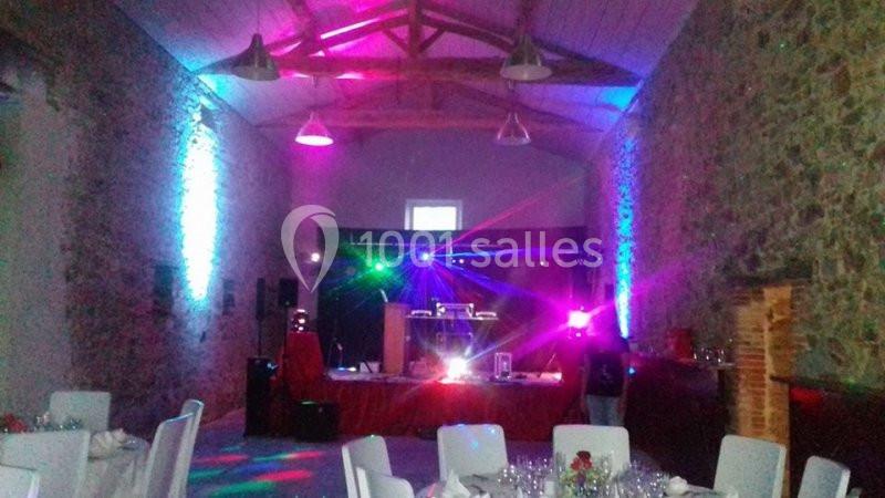 Salle en pierre aménagée pour un événement, avec éclairage coloré, tables dressées et espace DJ au fond.