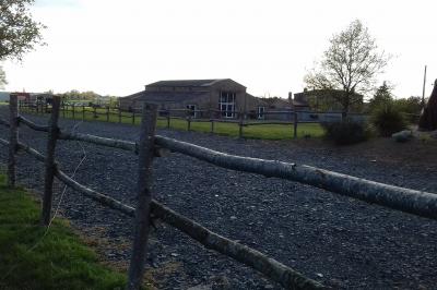 Chemin gravillonné bordé de barrières en bois menant à un bâtiment en pierre entouré de verdure.