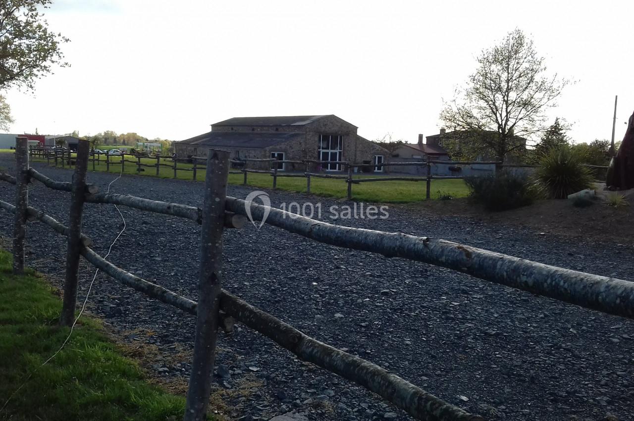Chemin gravillonné bordé de barrières en bois menant à un bâtiment en pierre entouré de verdure.