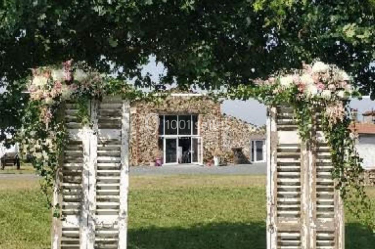 Deux volets en bois vieilli décorés de feuillage et de fleurs, formant une arche sur une pelouse devant un bâtiment en…