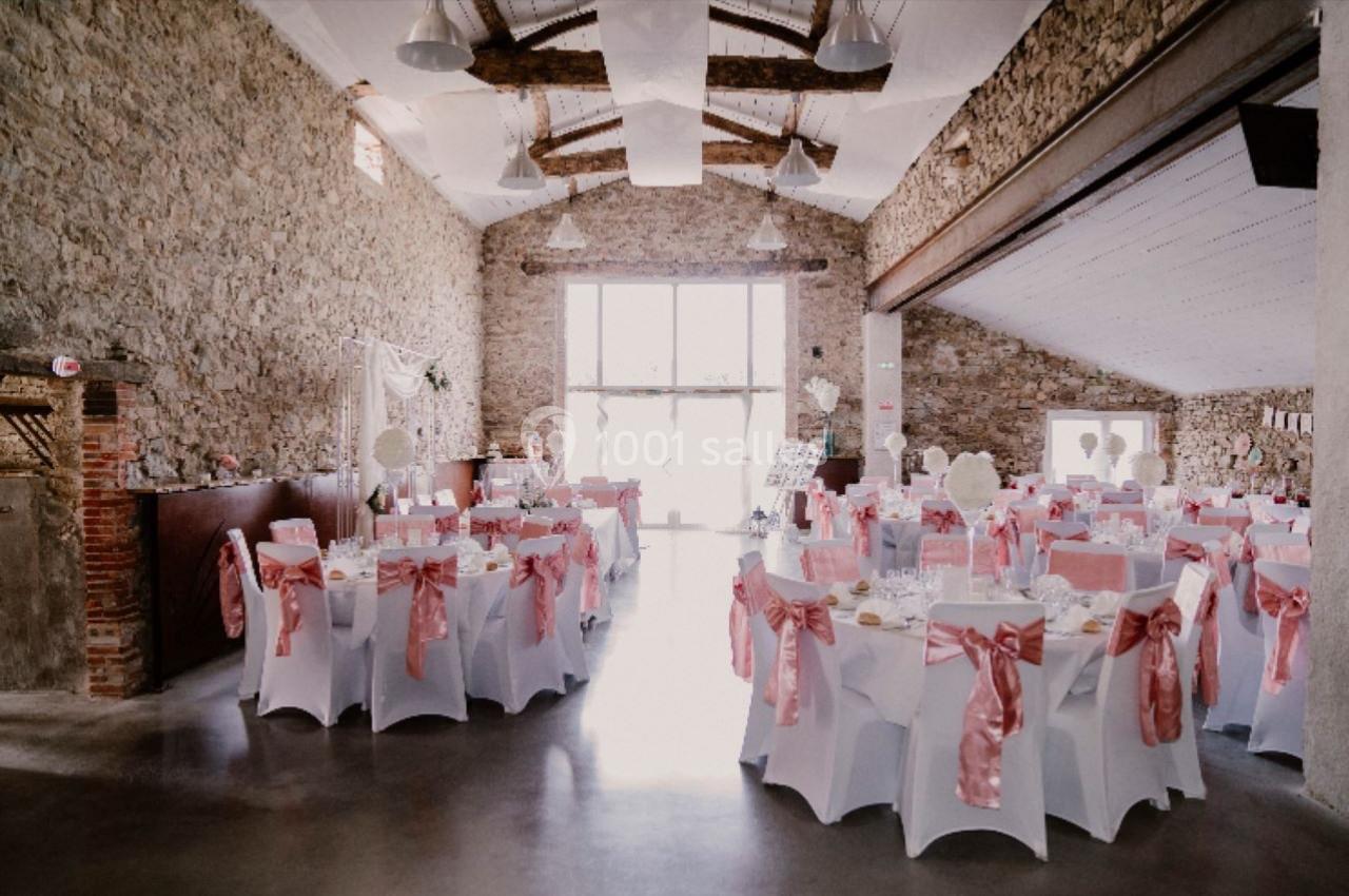 Salle de réception décorée avec des nappes blanches et des nœuds roses, éclairée par une grande baie vitrée.