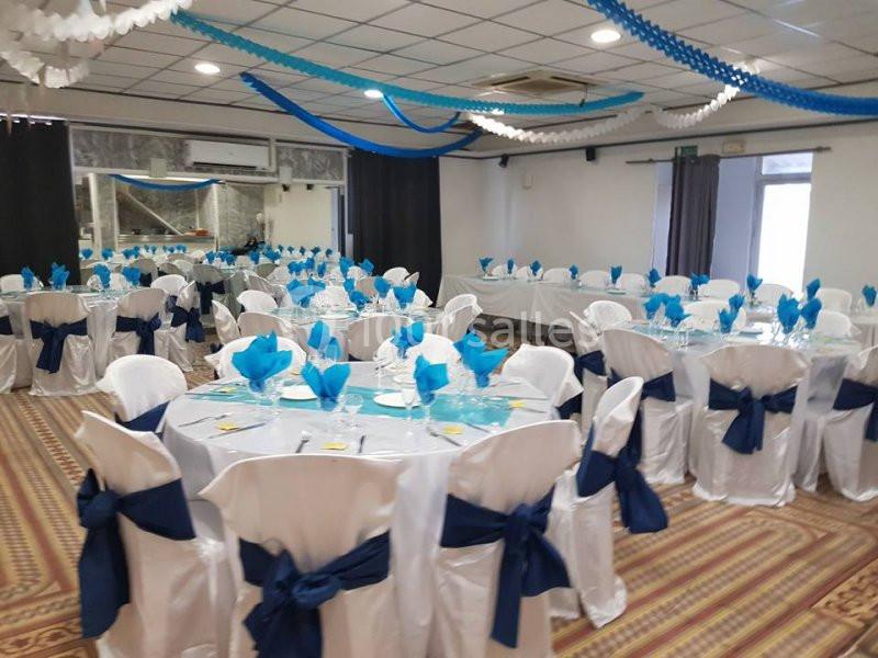 Salle de réception décorée avec des nappes blanches, des serviettes bleues et des rubans suspendus au plafond.