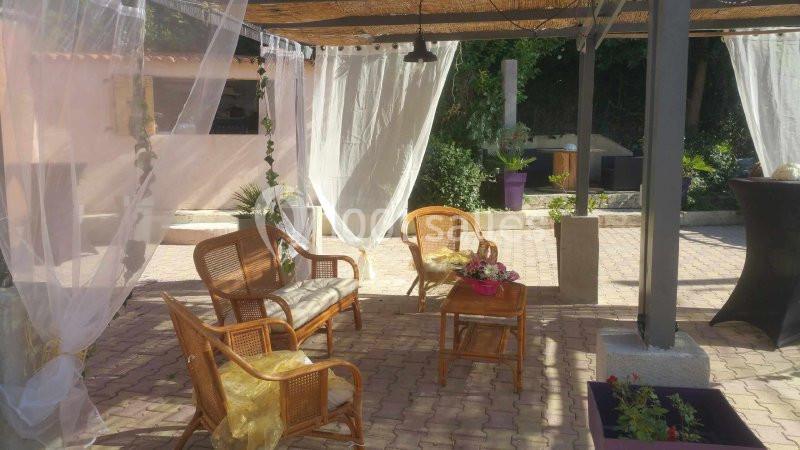 Espace extérieur aménagé avec des fauteuils en rotin, une table basse et des rideaux blancs sous une pergola.