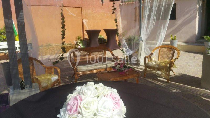 Espace extérieur aménagé avec des fauteuils en rotin, une table basse et des décorations florales sous des voilages.