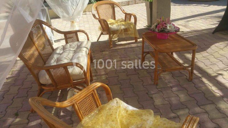 Salon de jardin en rotin avec coussins clairs, table basse décorée de fleurs, sur un sol pavé en extérieur.
