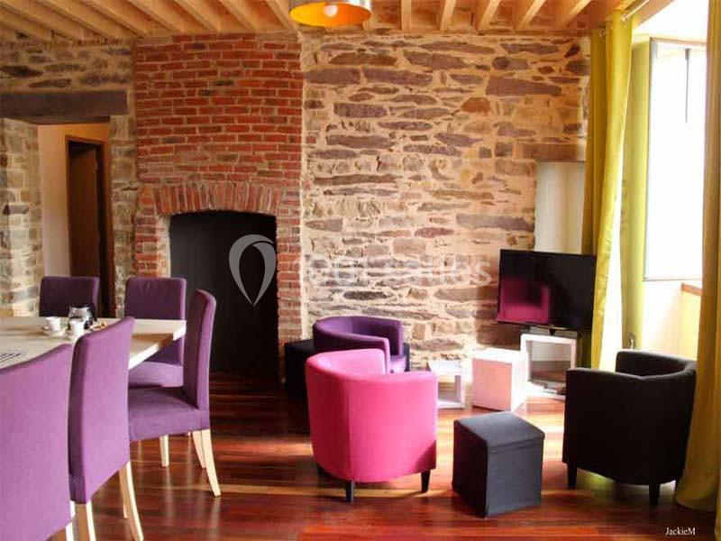 Salle de séjour avec murs en pierre, mobilier coloré, table à manger et télévision sur un meuble bas.