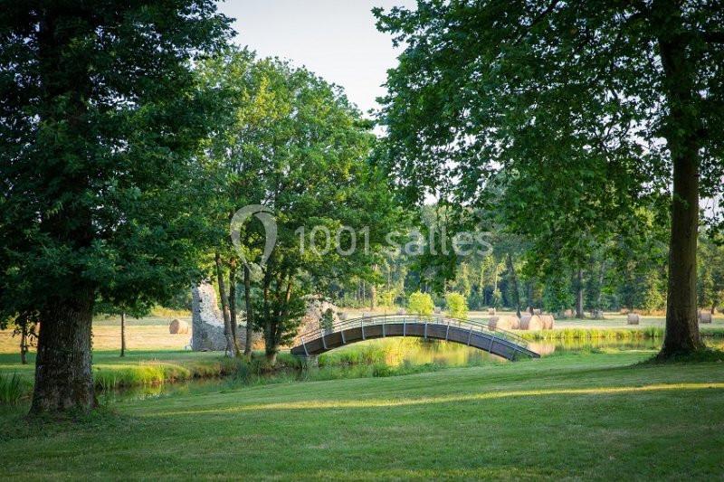 Petit pont en bois traversant un ruisseau, entouré de pelouses, d'arbres et de champs avec des bottes de foin.