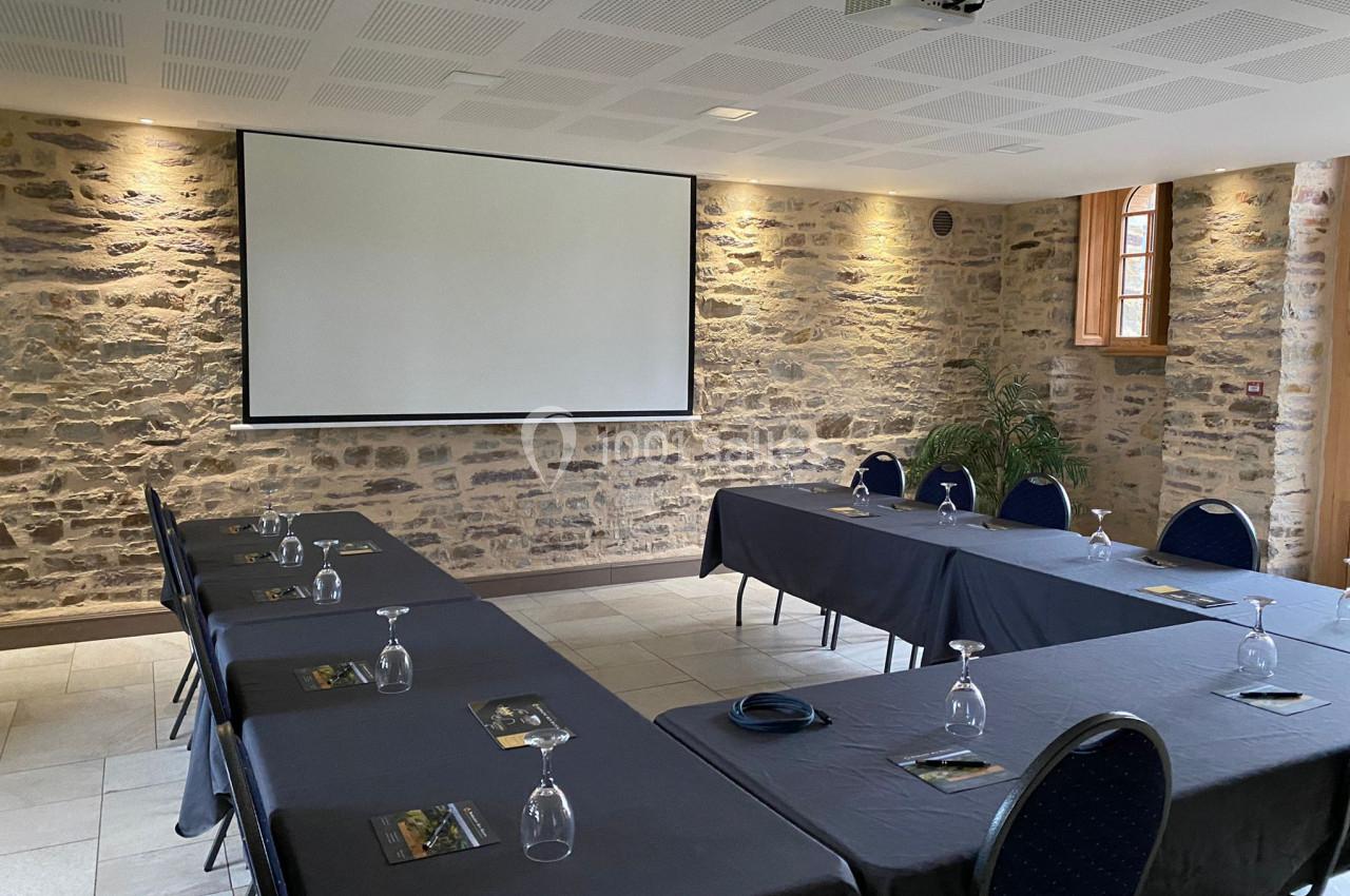 Salle de réunion avec murs en pierre, tables disposées en U, verres et documents posés, écran de projection au fond.