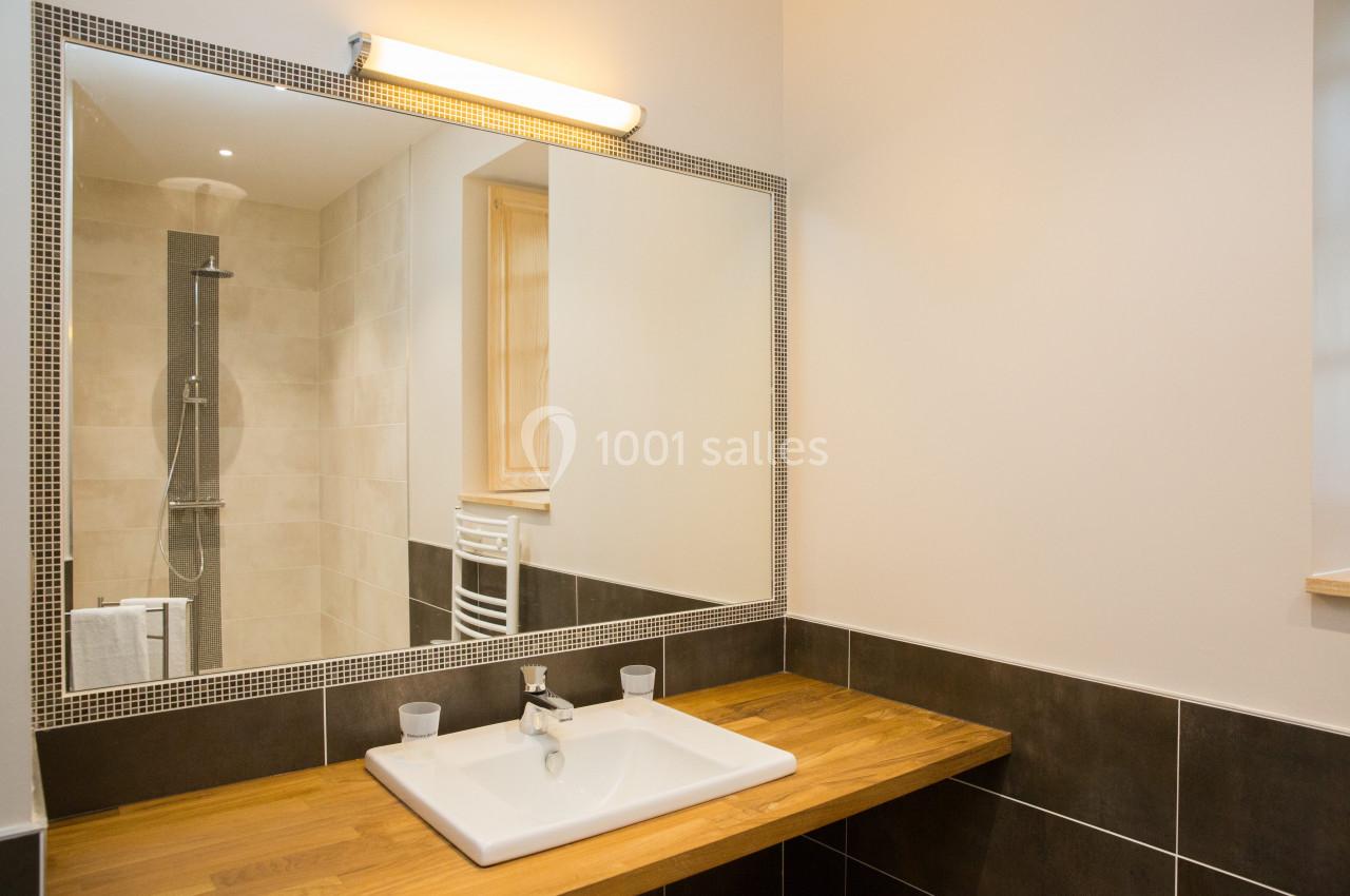 Lavabo avec plan de travail en bois, miroir éclairé, et douche en arrière-plan dans une salle de bain moderne.