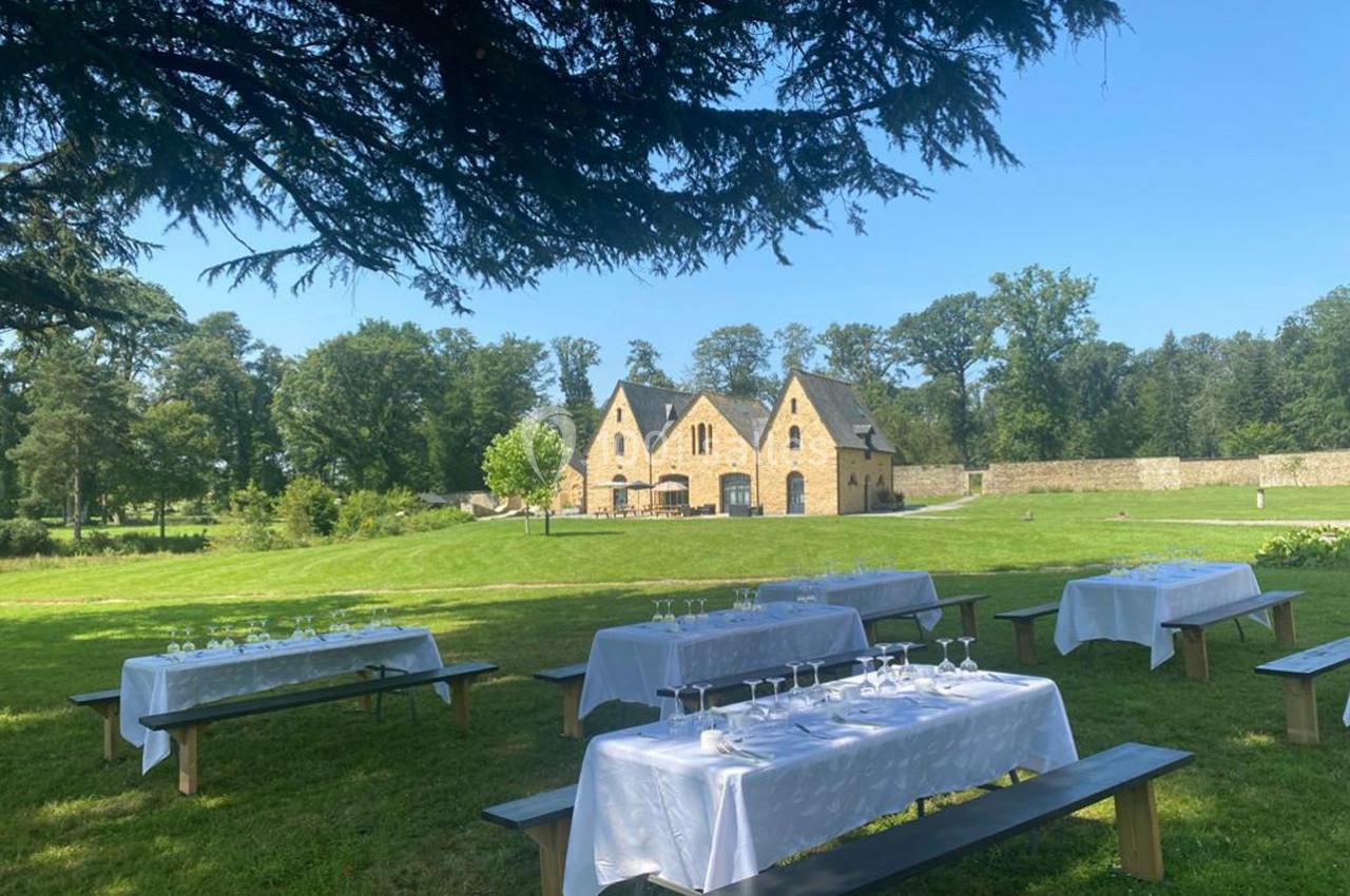 Tables dressées avec nappes blanches et verres sur des bancs en bois, disposées dans un parc devant un bâtiment en pierre.
