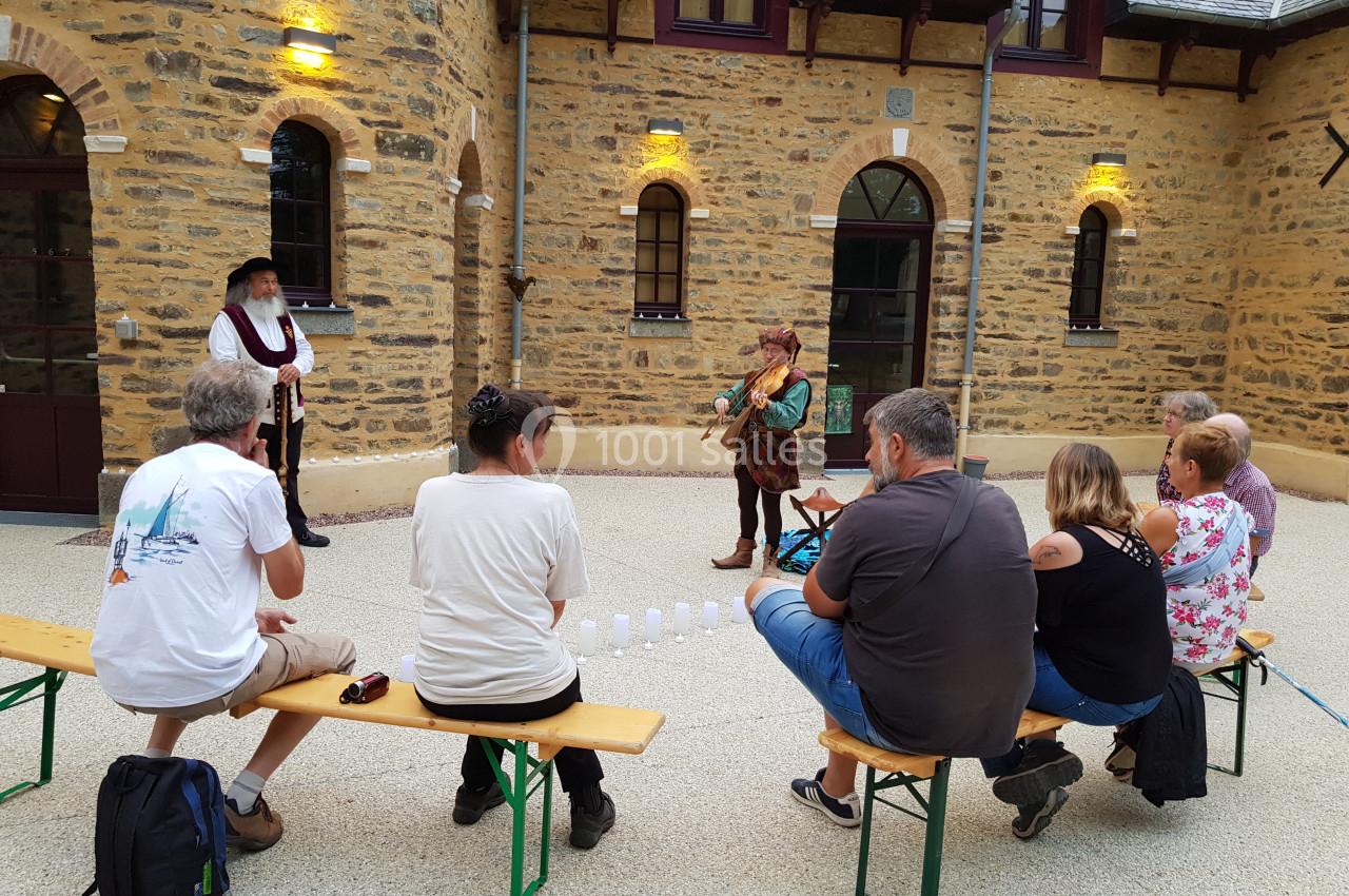 Un musicien en tenue médiévale joue de la flûte devant un petit groupe assis dans une cour en pierre.