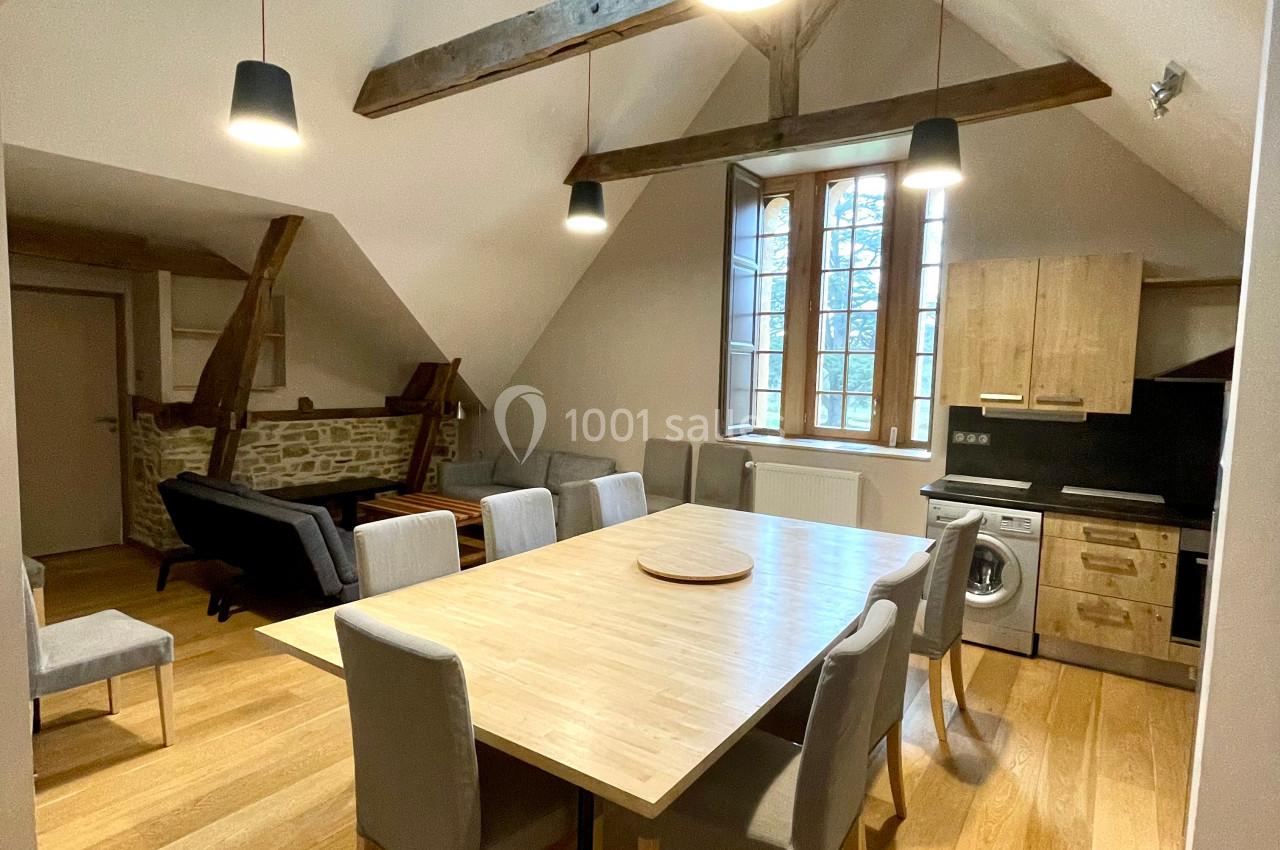 Salle à manger lumineuse avec table en bois, chaises grises, poutres apparentes et cuisine ouverte en arrière-plan.