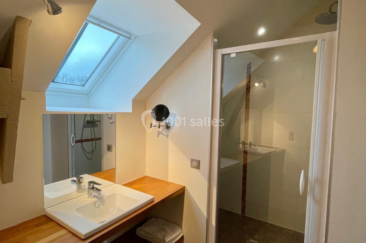 Salle de bain mansardée avec lavabo sur plan en bois, miroir, douche vitrée et fenêtre de toit.