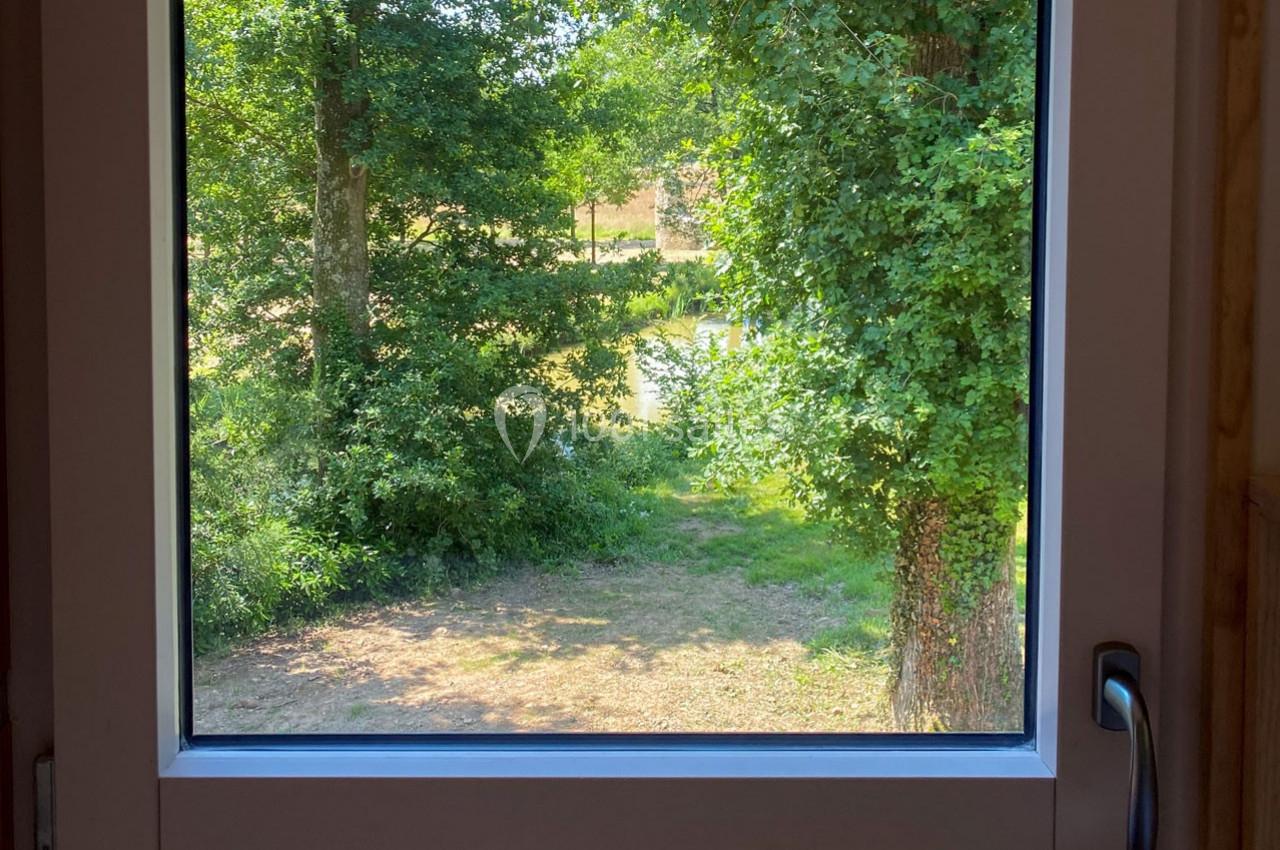 Vue depuis une fenêtre ouverte sur un jardin verdoyant avec des arbres et un chemin ensoleillé.