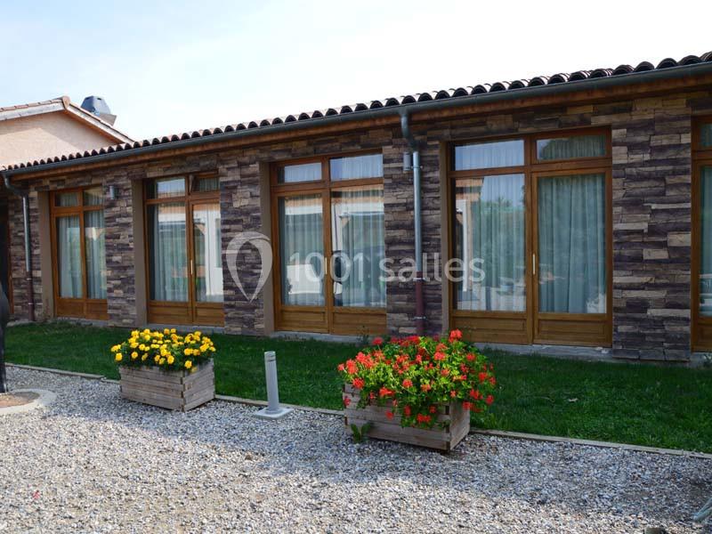 Façade d'un bâtiment en pierre avec de grandes baies vitrées, entouré de jardinières fleuries et d'une allée gravillonnée.
