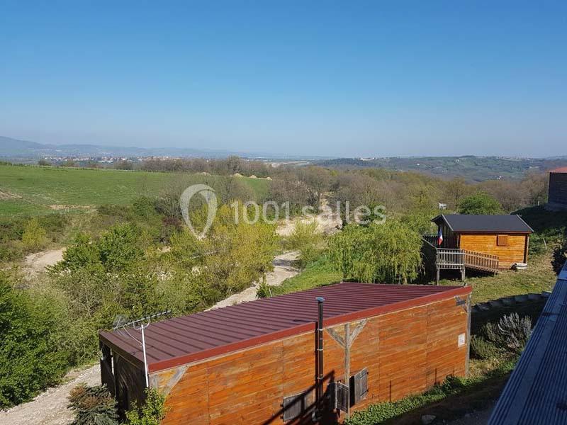 Vue sur des cabanes en bois entourées de végétation, avec un paysage vallonné et un ciel dégagé en arrière-plan.