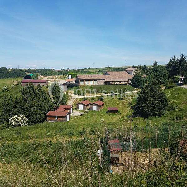 Vue d'un paysage rural avec des bâtiments en pierre, des arbres et des enclos sur un terrain vallonné.