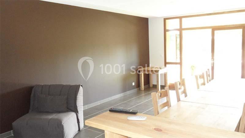 Salle lumineuse avec un mur marron, une table en bois entourée de chaises, un fauteuil gris et une télécommande sur la table.