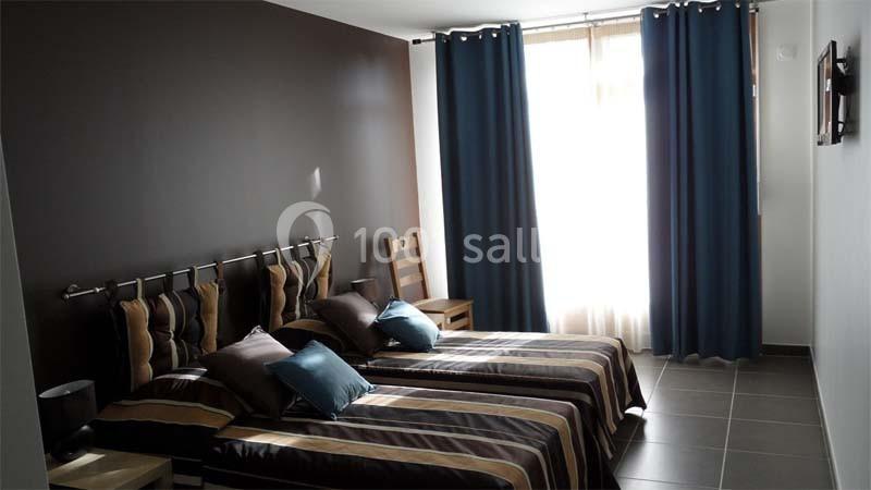 Chambre avec deux lits simples, murs sombres, rideaux bleus, lumière naturelle entrant par une grande fenêtre.