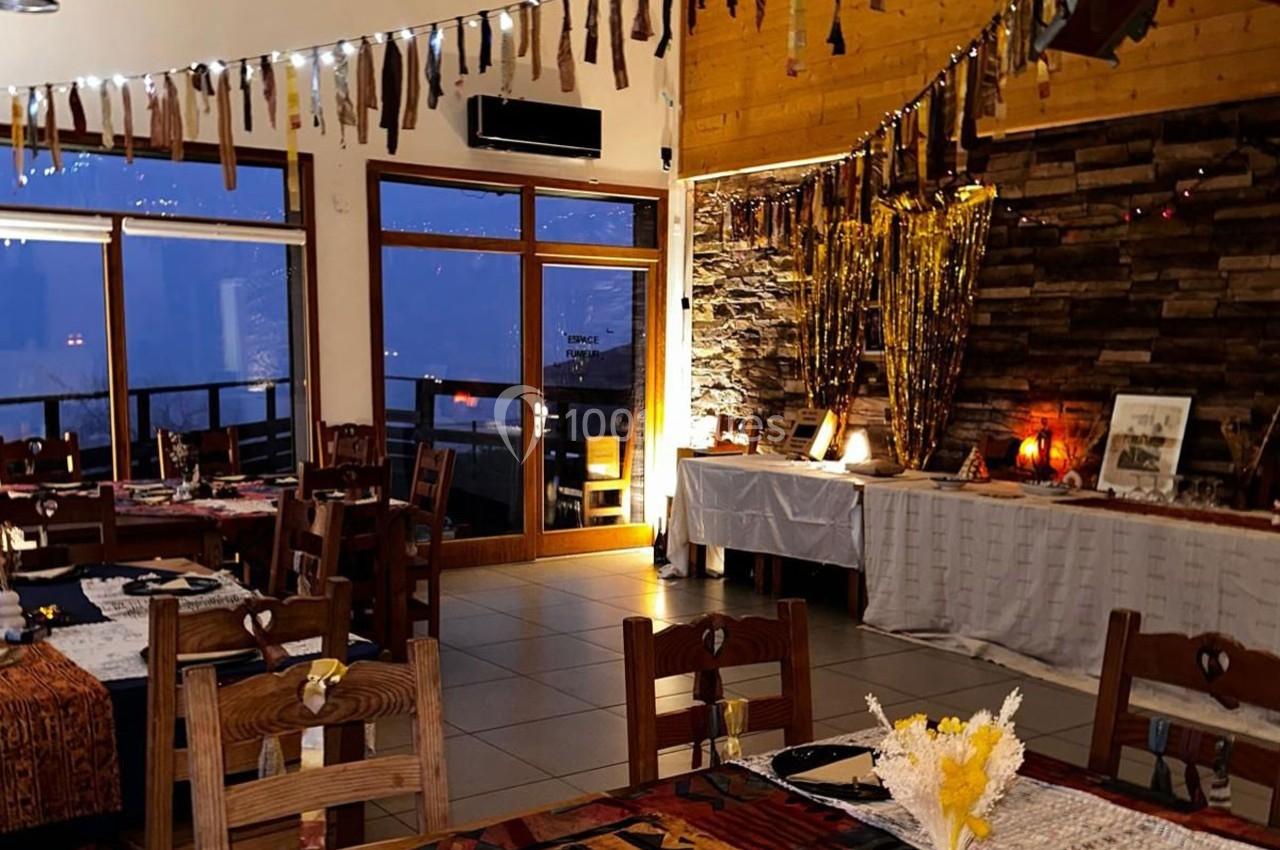 Salle de restaurant chaleureuse avec tables décorées, éclairage tamisé et vue sur l'extérieur à travers de grandes baies…