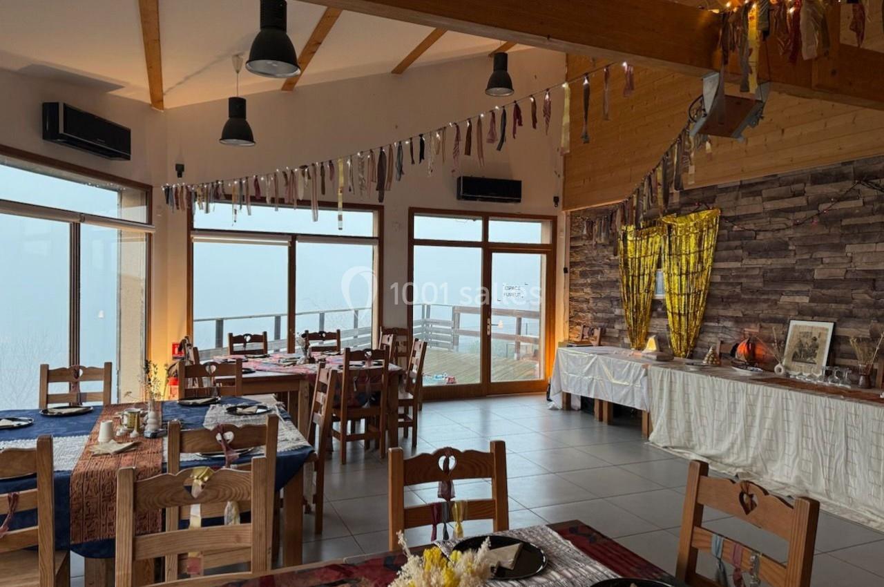 Salle de restaurant lumineuse avec grandes baies vitrées, tables dressées et décorations suspendues.