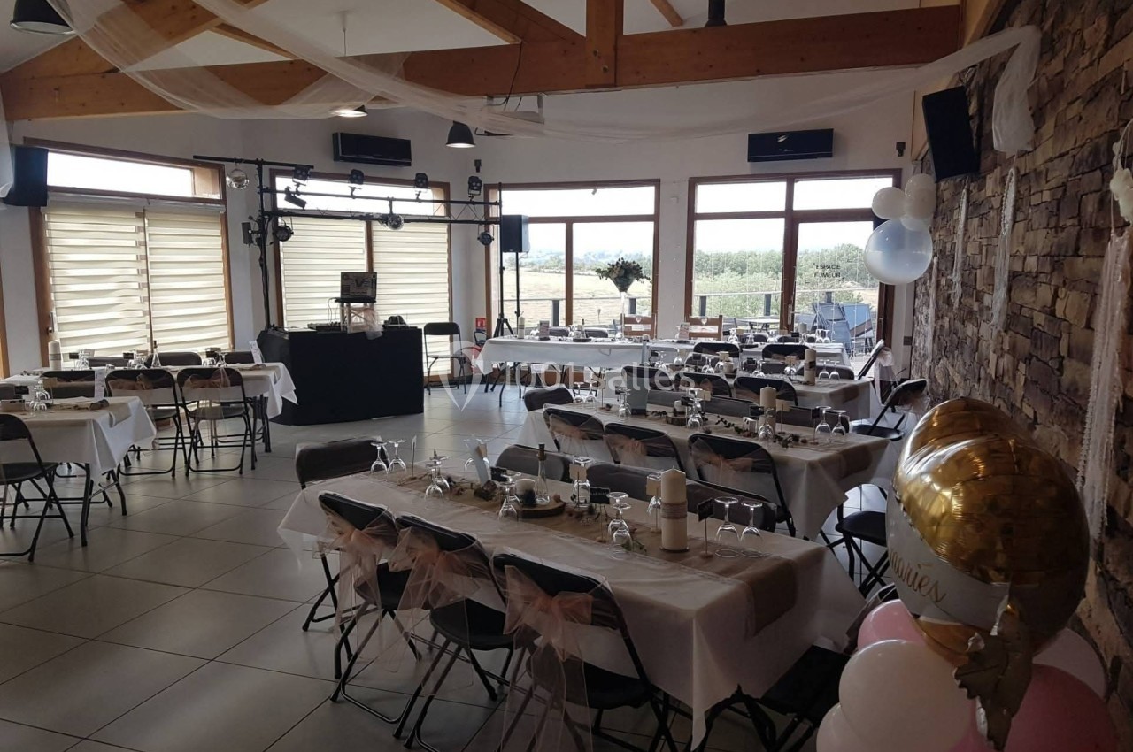 Salle décorée pour un événement avec tables dressées, chaises ornées de nœuds roses et ballons près d'un mur en pierre.