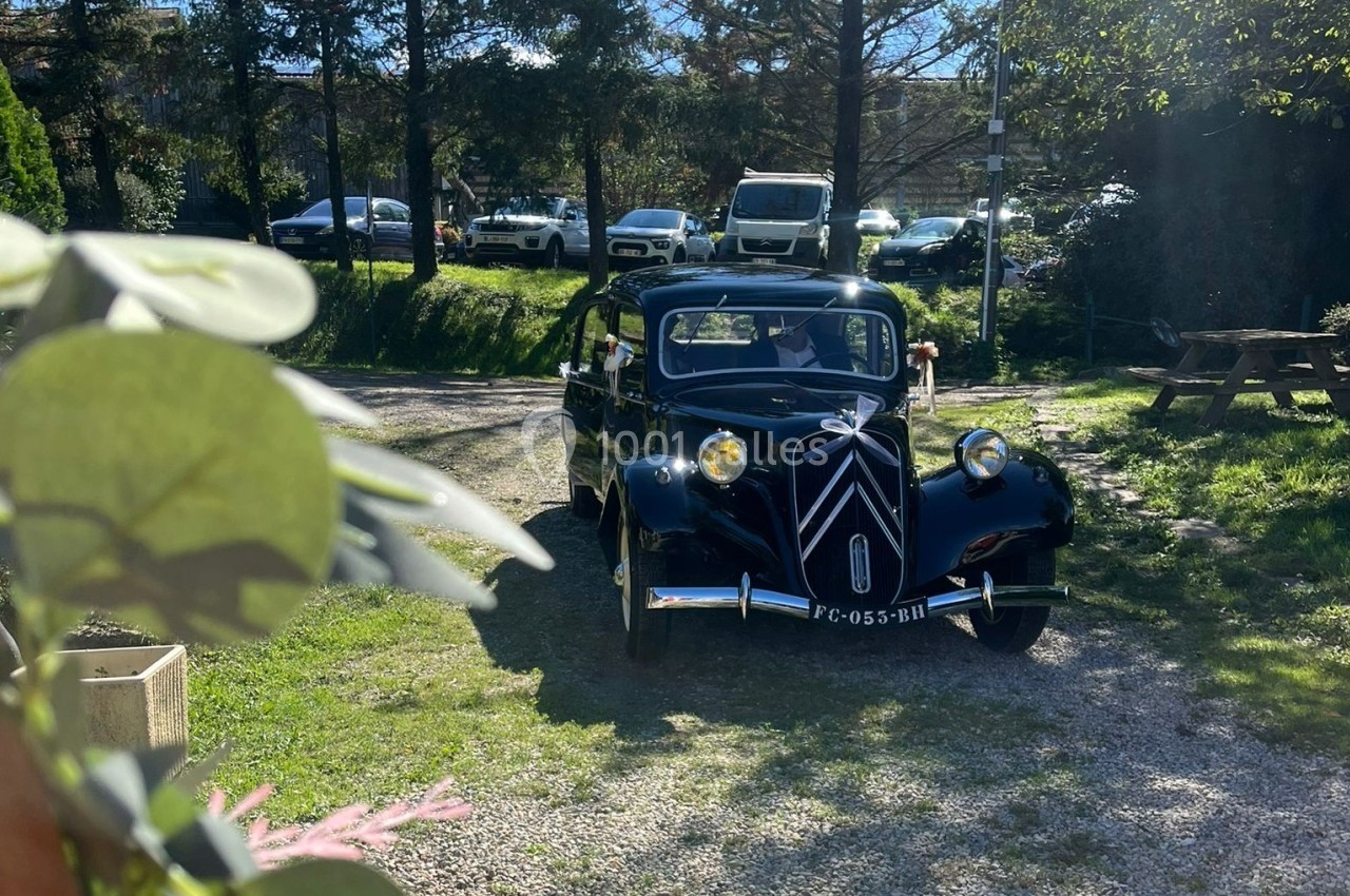 Voiture ancienne noire circulant sur un chemin gravillonné entouré de verdure et de véhicules stationnés.