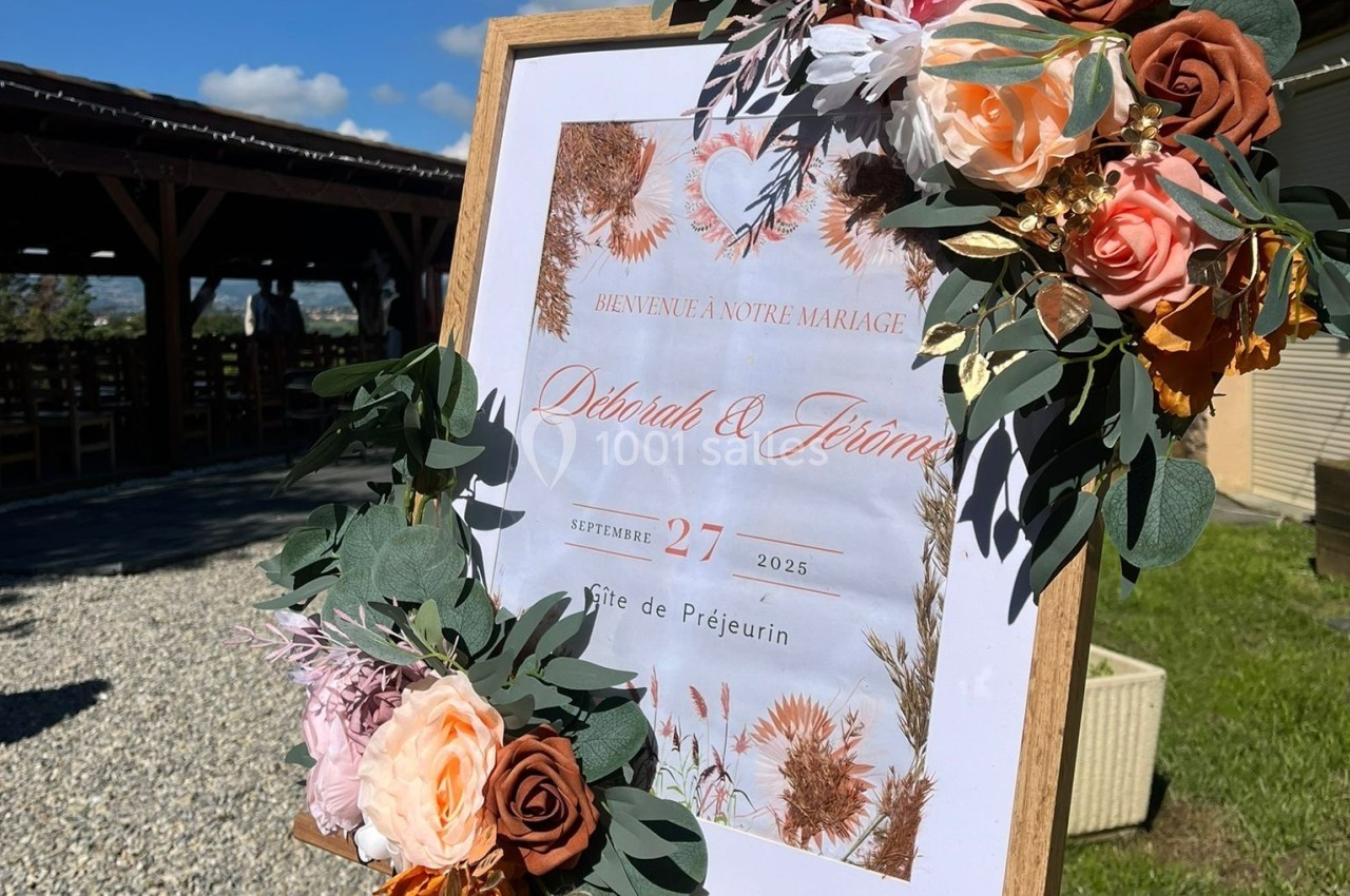 Panneau de bienvenue pour un mariage, décoré de fleurs et feuillages, placé à l'entrée d'un espace extérieur.