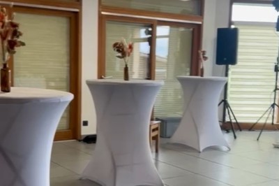 Tables hautes recouvertes de nappes blanches, décorées de bouquets, dans une salle lumineuse avec baies vitrées.