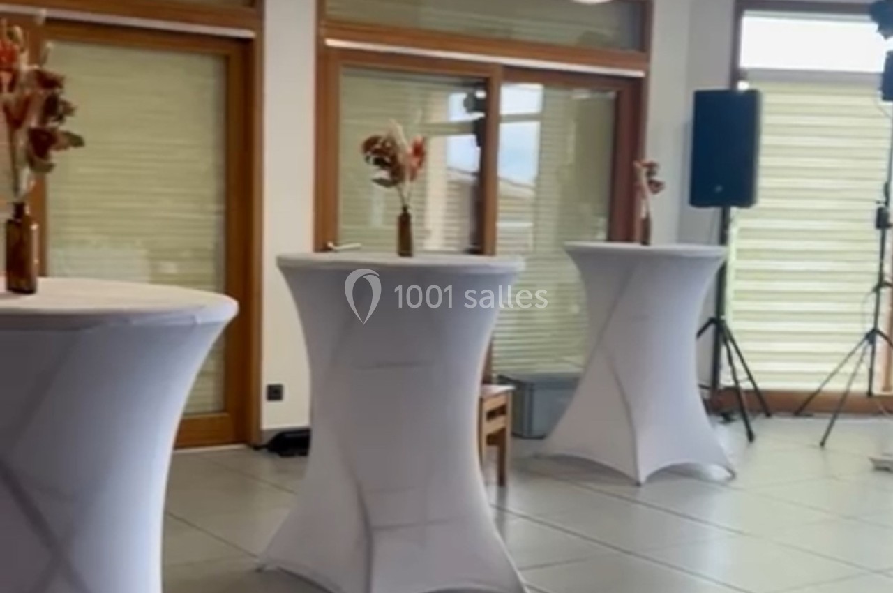 Tables hautes recouvertes de nappes blanches, décorées de bouquets, dans une salle lumineuse avec baies vitrées.