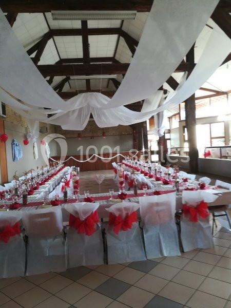 Salle de réception décorée avec des tables en U, nappes blanches, chaises ornées de nœuds rouges et voilages suspendus.