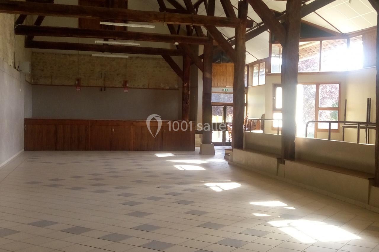 Grande salle lumineuse avec charpente en bois apparente, sol carrelé et murs clairs, éclairée par des fenêtres.