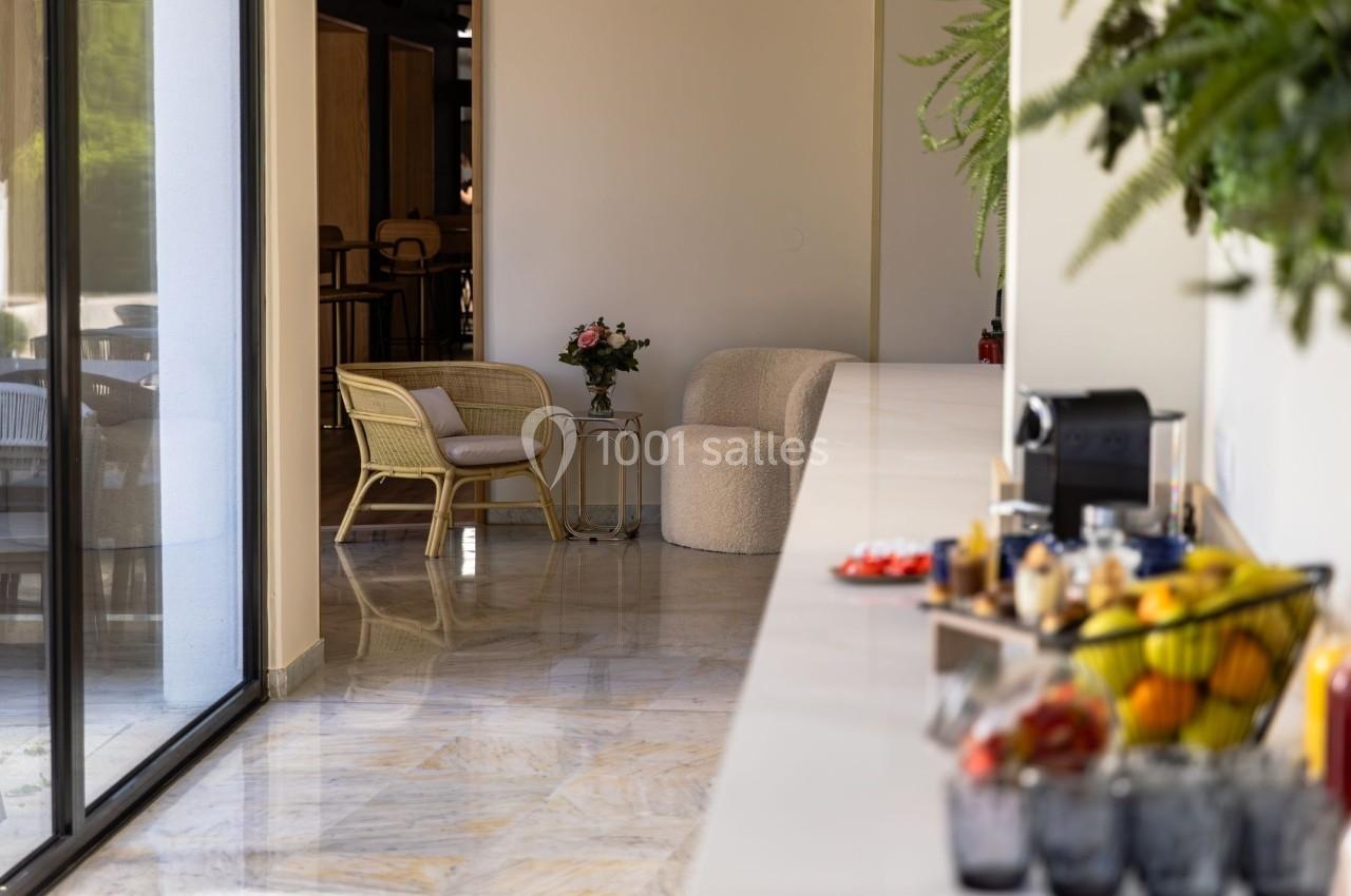Espace lumineux avec fauteuils, table basse et plantes, vue sur un comptoir garni de fruits et boissons.