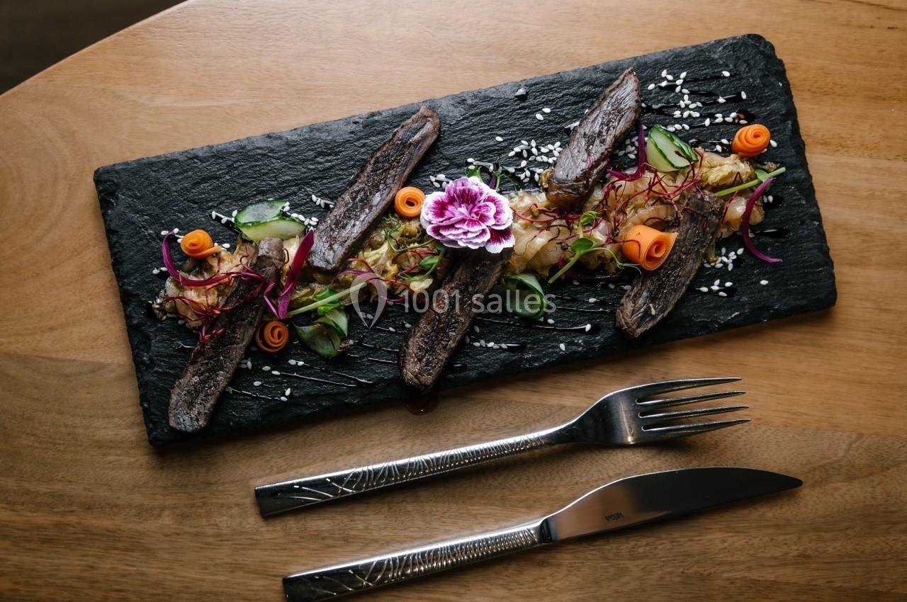 Assiette en ardoise garnie de viande grillée, légumes décoratifs et fleurs comestibles, avec couteau et fourchette.