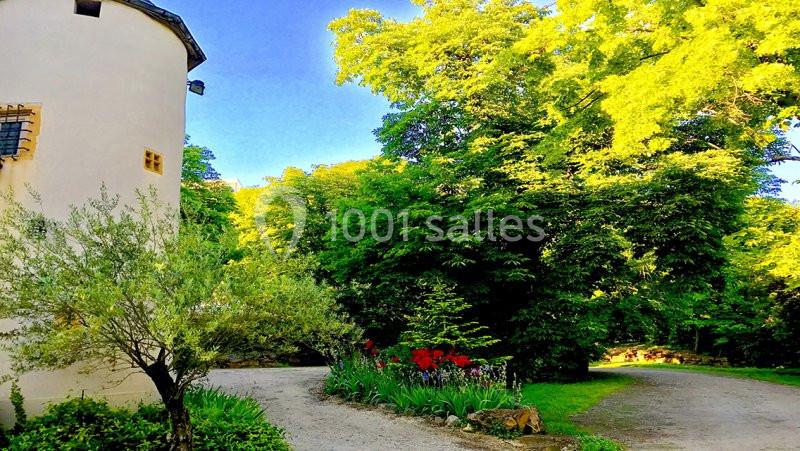 Chemin bordé de verdure et de fleurs rouges, près d'un bâtiment clair et entouré d'arbres sous un ciel bleu.