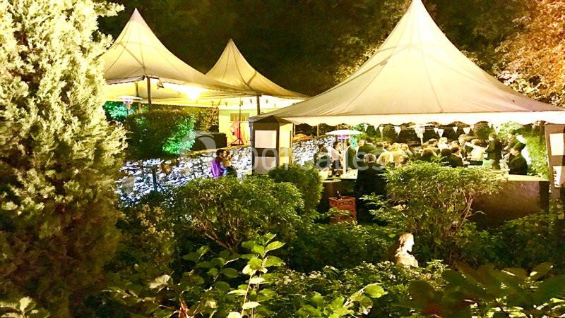 Des invités rassemblés sous des tentes blanches dans un jardin éclairé en soirée, entouré de végétation dense.