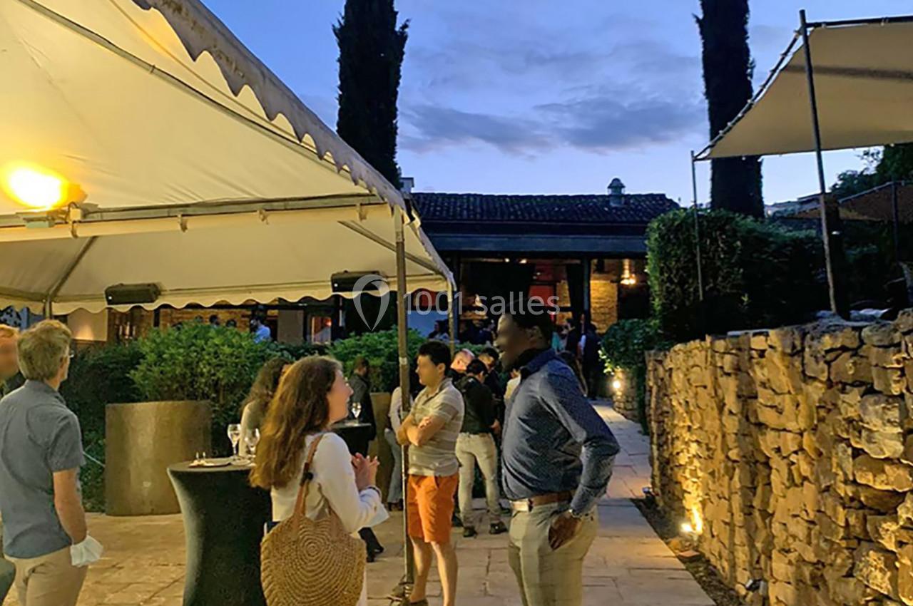 Personnes discutant dans une cour extérieure en soirée, éclairée par des lampes et entourée de murs en pierre.