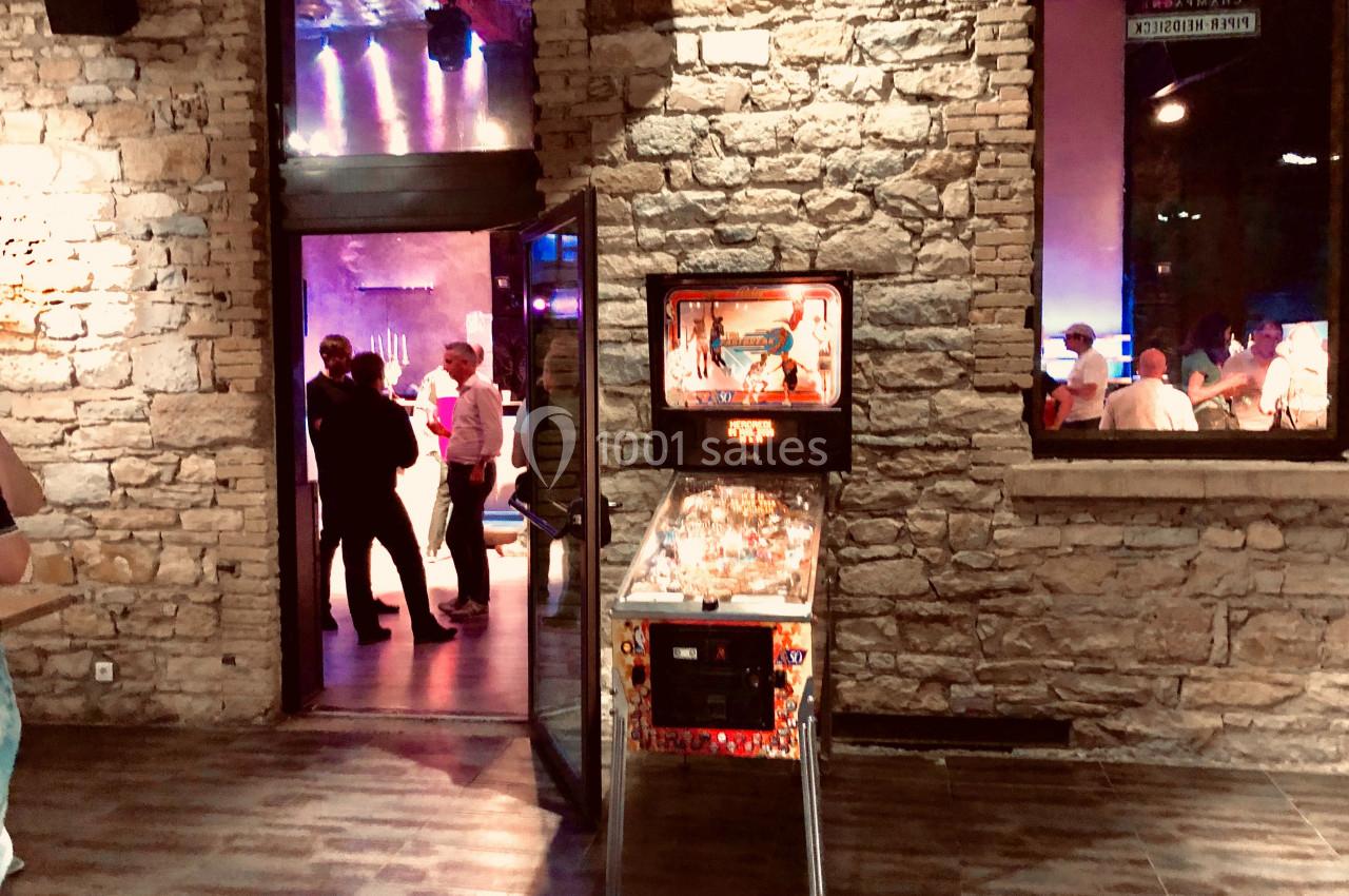 Un flipper éclairé dans un espace en pierre, avec des personnes discutant dans une pièce adjacente animée.