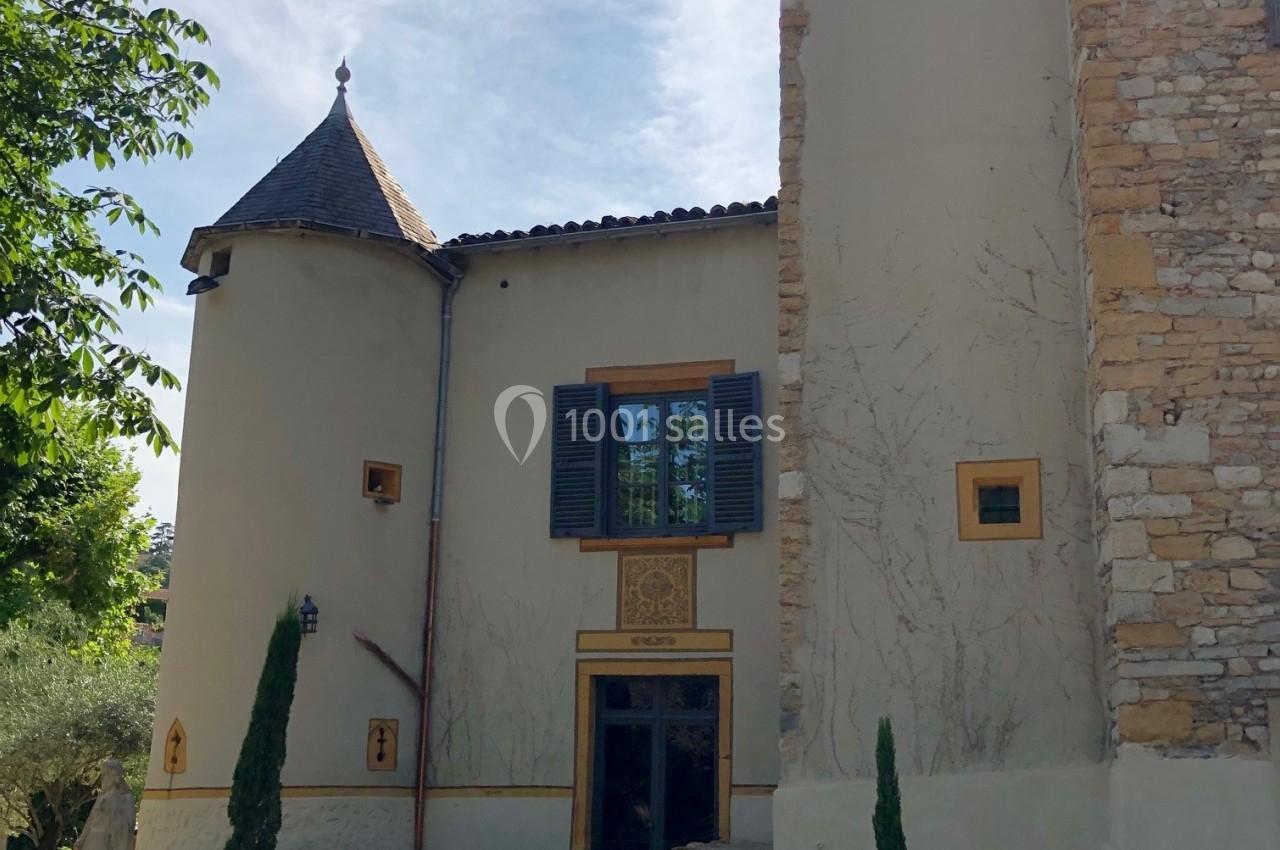 Façade d'une maison en pierre avec deux tours, fenêtres à volets et terrasse aménagée avec mobilier d'extérieur.