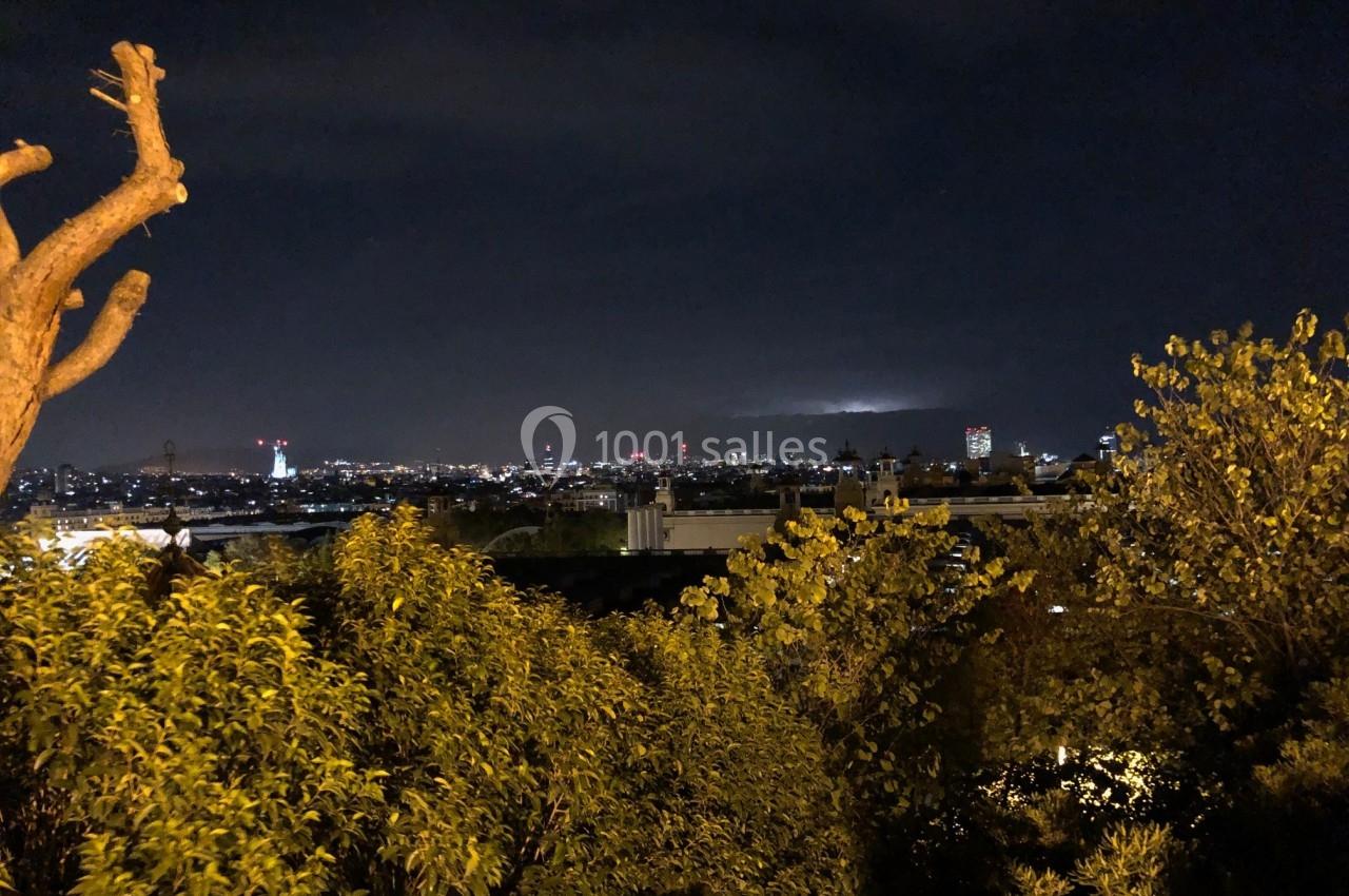 Vue nocturne d'une ville illuminée à l'horizon, entourée d'arbres et de végétation au premier plan.