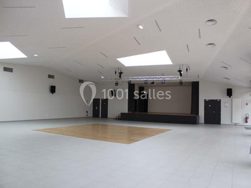 Salle polyvalente vide avec scène, parquet central, sol carrelé et éclairage naturel provenant de puits de lumière.