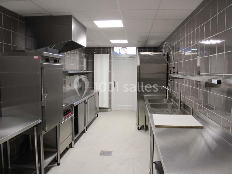 Cuisine professionnelle équipée avec plans de travail en inox, éviers, four et hotte dans un espace carrelé.