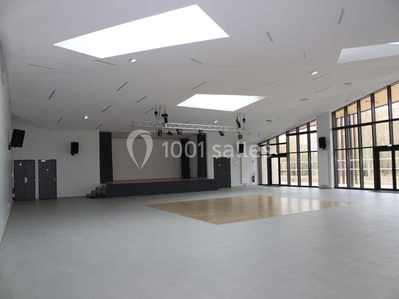Salle polyvalente lumineuse avec scène, parquet central, grandes baies vitrées et éclairage moderne.