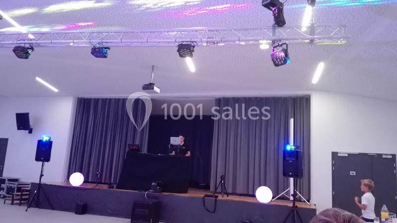 Une scène avec un DJ derrière une table, éclairée par des lumières colorées, dans une salle équipée de haut-parleurs.