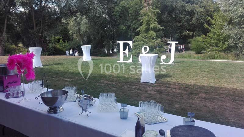 Table dressée en extérieur avec verres et accessoires de réception, grand jardin en arrière-plan avec lettres décoratives ’E…