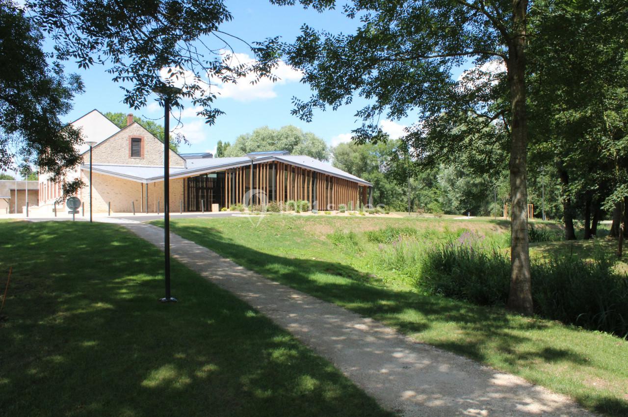 Bâtiment moderne en pierre et bois entouré de verdure, avec un chemin menant à l'entrée sous un ciel ensoleillé.