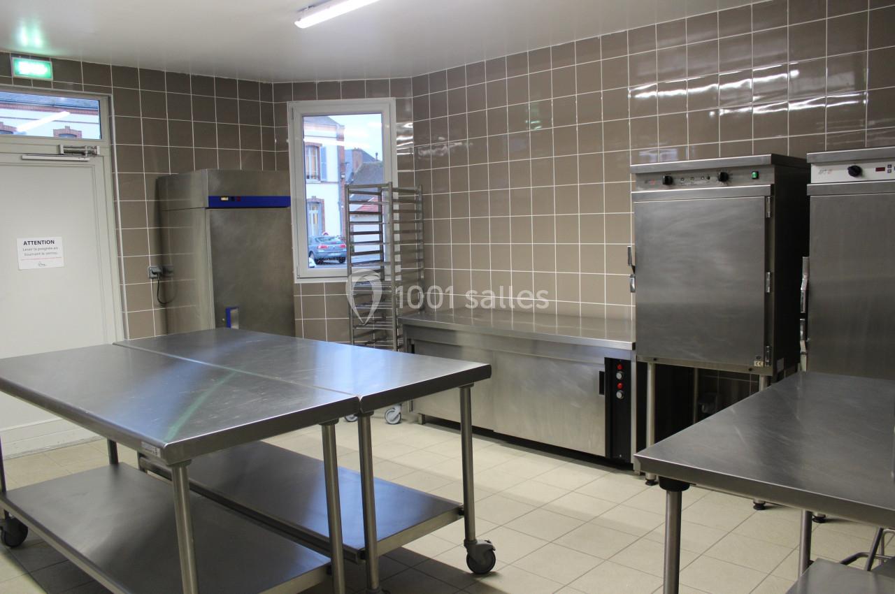 Cuisine professionnelle avec plans de travail en inox, étagères, fours et carrelage beige sur les murs.