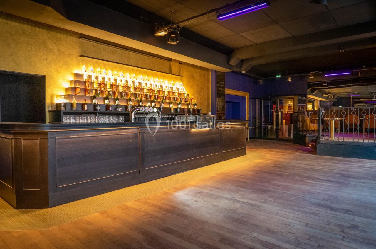 Bar en bois sombre avec étagères illuminées de bouteilles, situé dans une grande salle au sol en parquet.