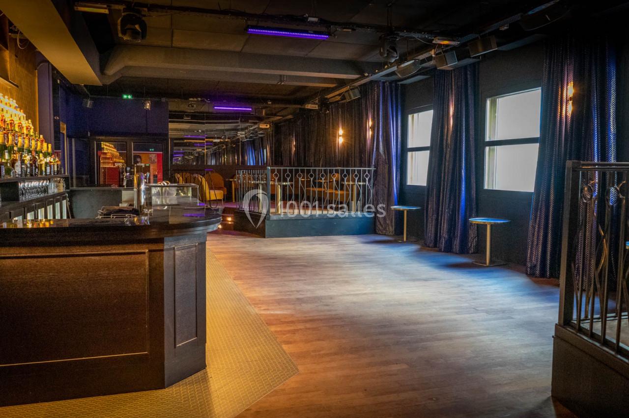 Salle de bar avec comptoir en bois sombre, éclairage tamisé et rideaux bleus, espace ouvert avec quelques tabourets.