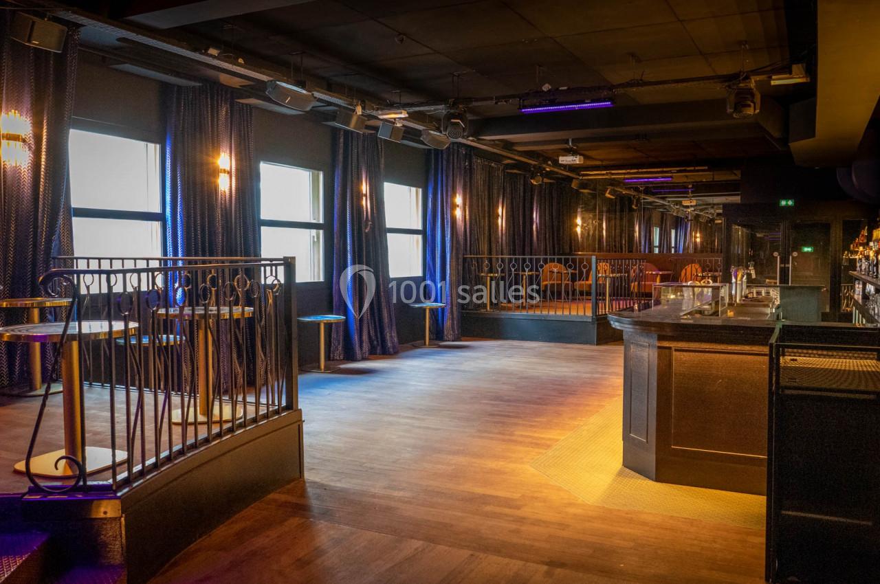 Salle de bar moderne avec éclairage tamisé, comptoir à droite, tables hautes et rideaux sombres en arrière-plan.